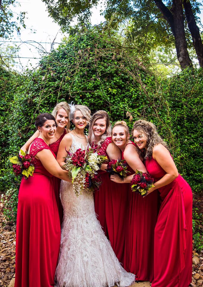 bride smiling holding flowers, smiling with her bridemaids wearing red dresses.
